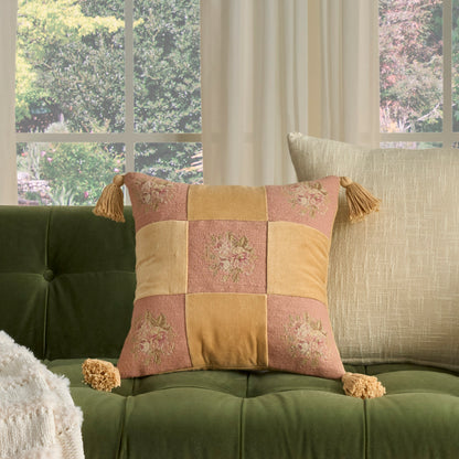 Patchwork decorative pillow in pink and mustard yellow with floral embroidery and corner tassels on a green tufted sofa.