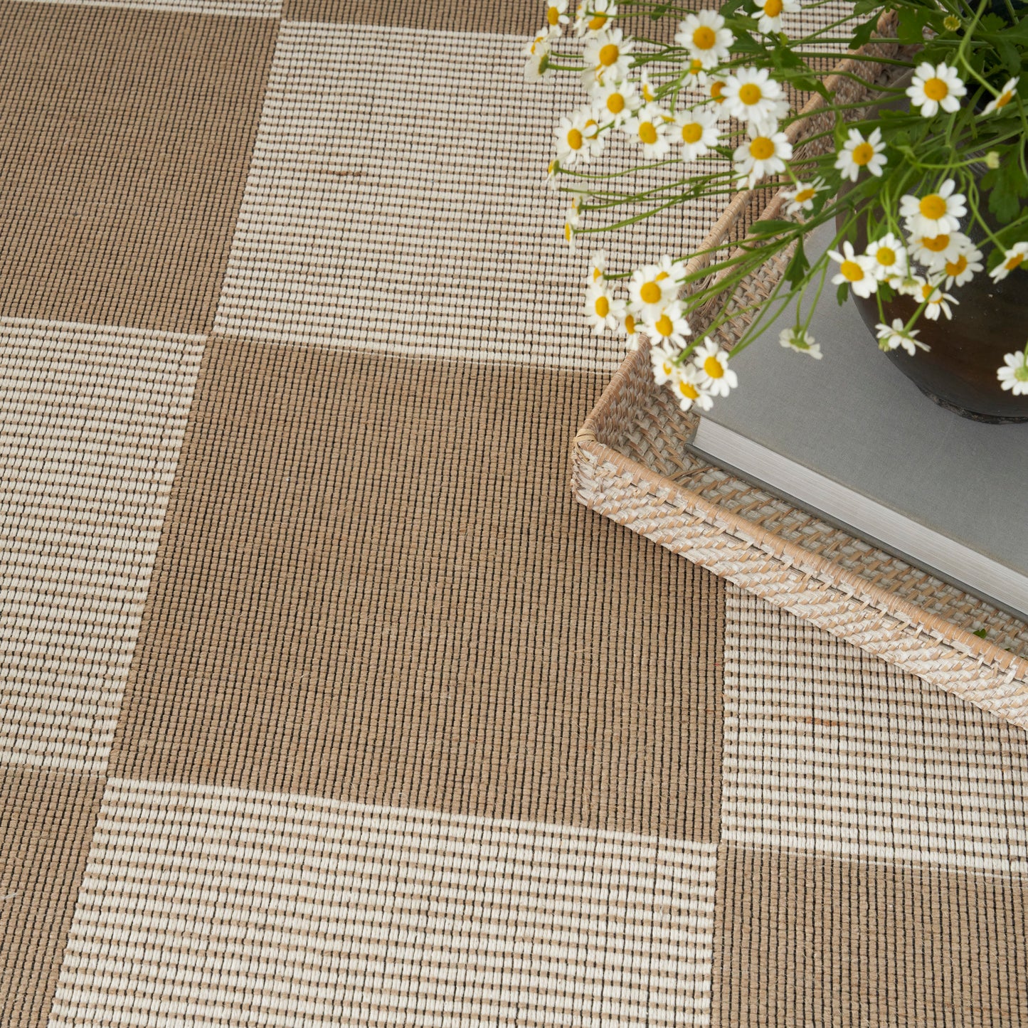 Brown and beige checkered woven rug with a wicker tray and a bouquet of white daisies.