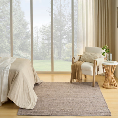 Neutral bedroom with beige bedspread, taupe woven rug, light-wood chair and round side table with plant by large windows.
