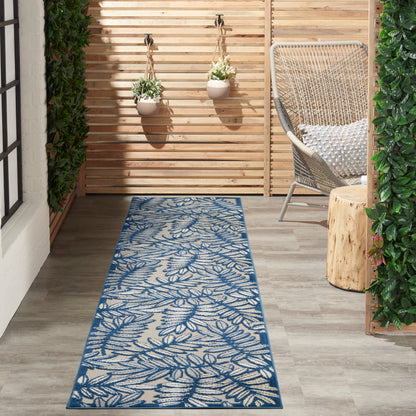 Outdoor runner rug with blue botanical pattern on beige background, on a wooden deck & beside a wicker chair & hanging planters.