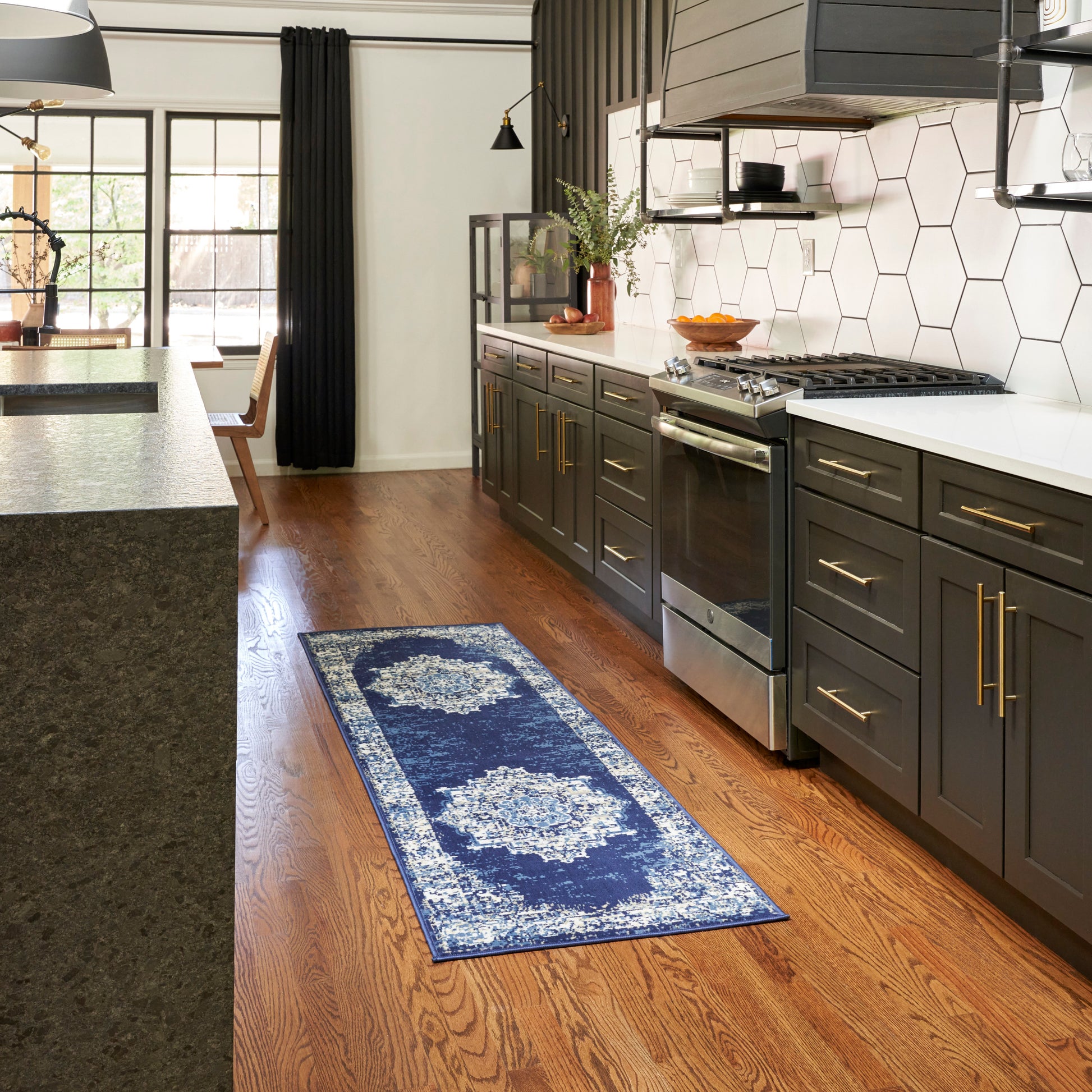 Blue patterned runner rug on hardwood floor beside gray cabinets, white hex tile backsplash, stainless gas range in a modern kitchen.