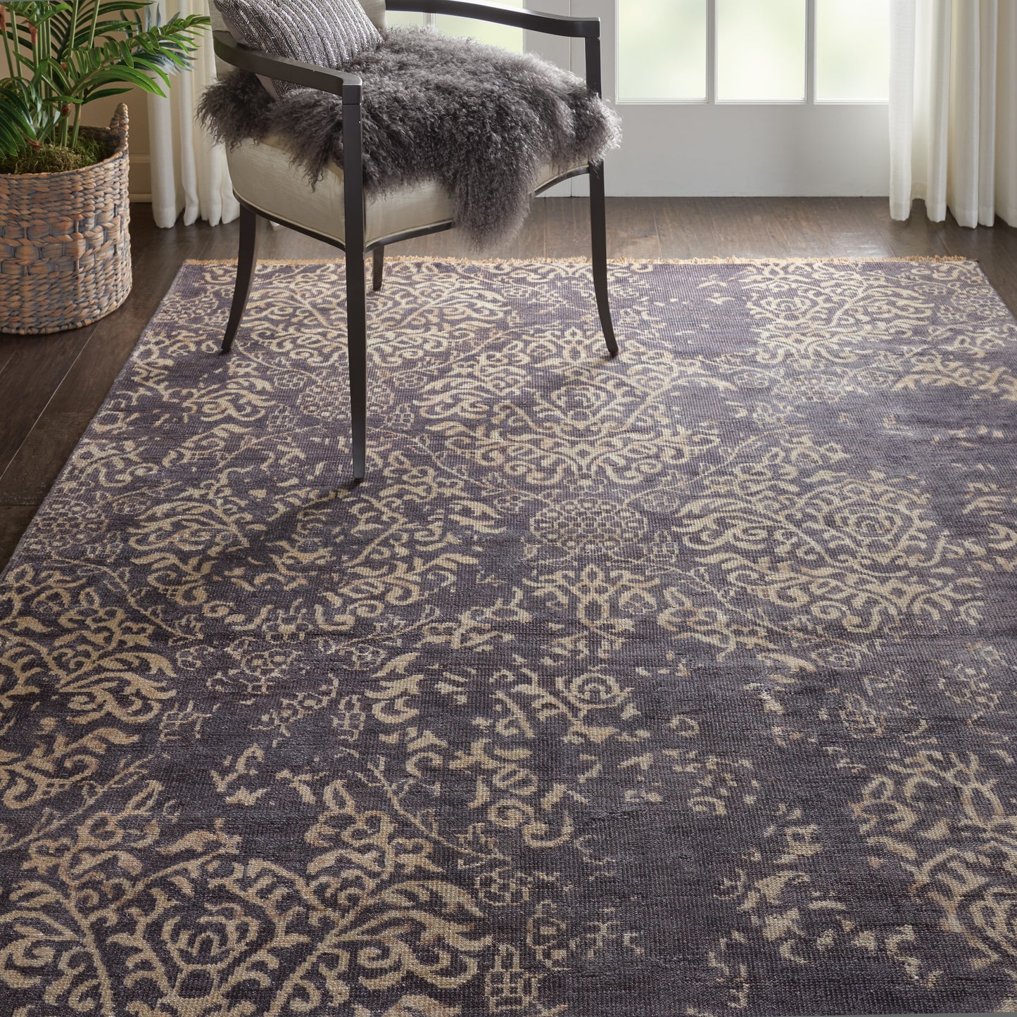 Charcoal area rug with beige ornate floral pattern in a bright living room with a metal-framed chair and gray faux fur throw.