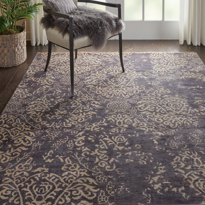 Charcoal area rug with beige ornate floral pattern in a bright living room with a metal-framed chair and gray faux fur throw.