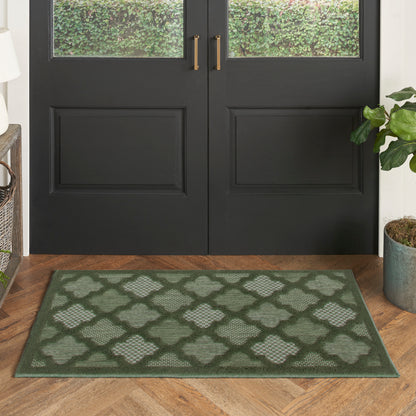 Green lattice-pattern rug on wood floor in front of double black doors with brass handles; potted plant nearby.