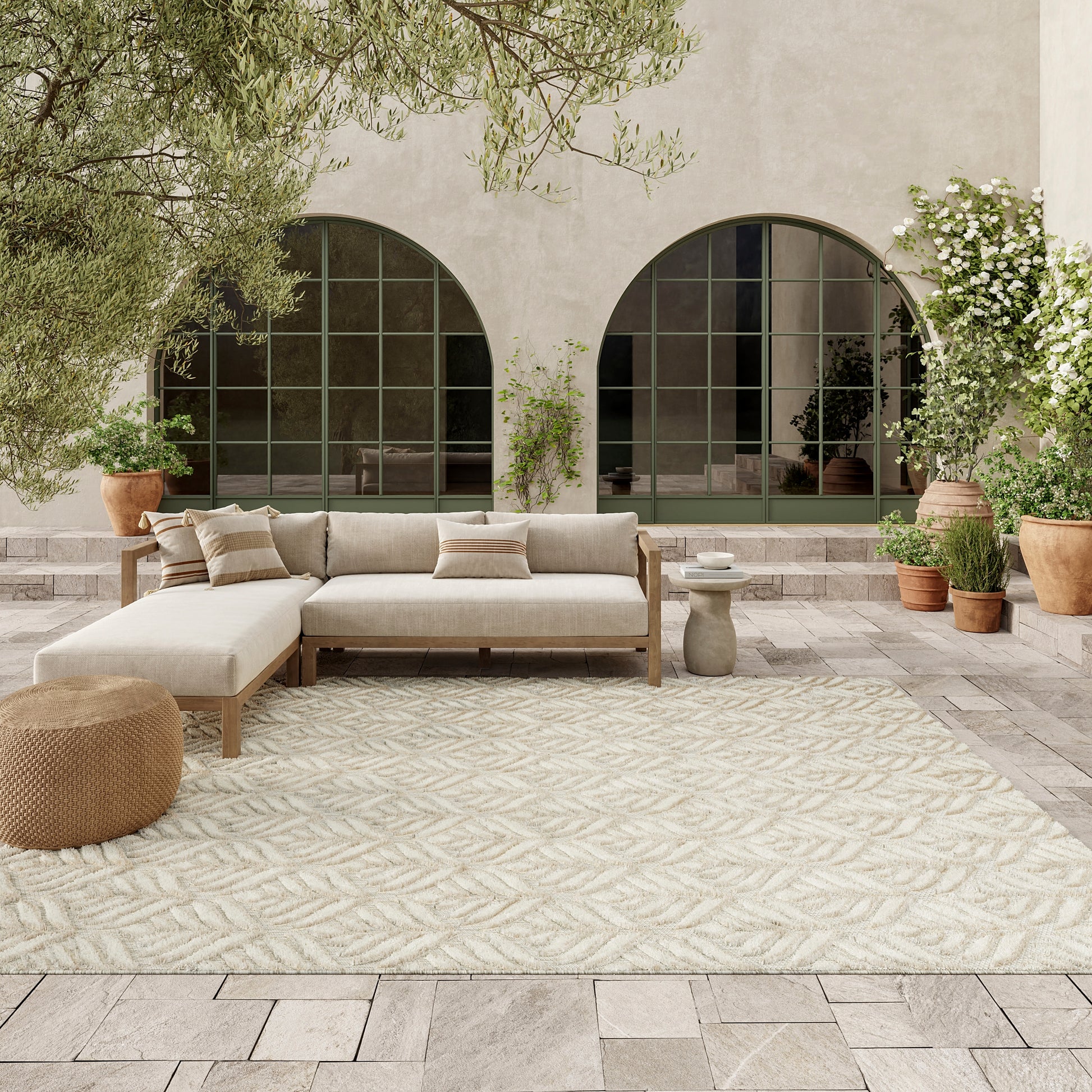 Wood-framed beige fabric sectional on a cream leaf-pattern rug, arched green-framed doors, and potted greenery.