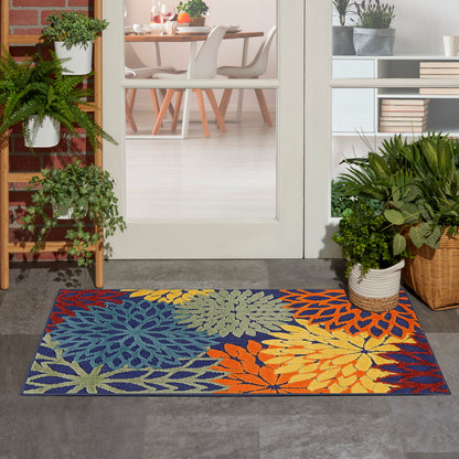 Colorful tropical floral doormat in blue, yellow, orange and green; rectangular outdoor entry rug by glass door with plants.