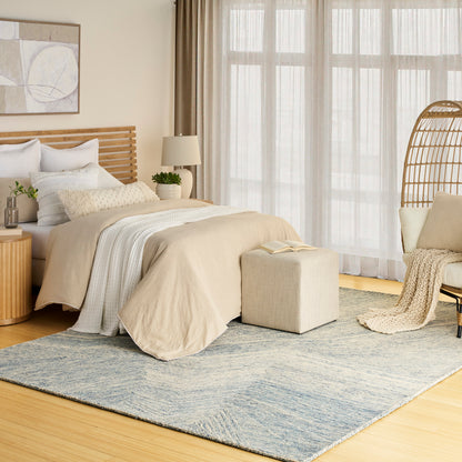 Neutral bedroom with beige bedding, white pillows, wooden slat headboard, blue rug, ottoman, wicker chair, sheer curtains.