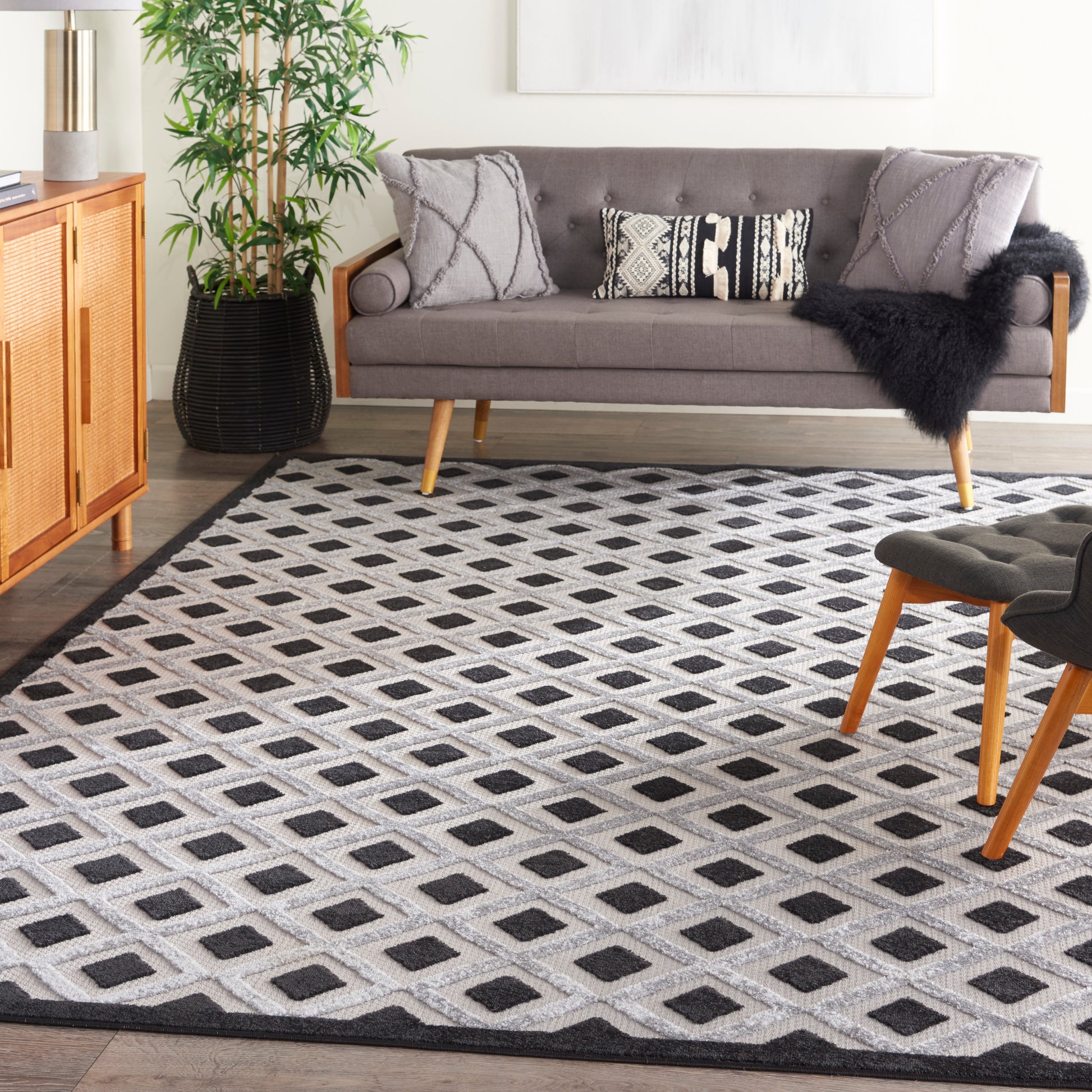 Living room with mid-century gray sofa, black-and-white diamond rug, wooden cabinet, potted plant & light-wood legs.