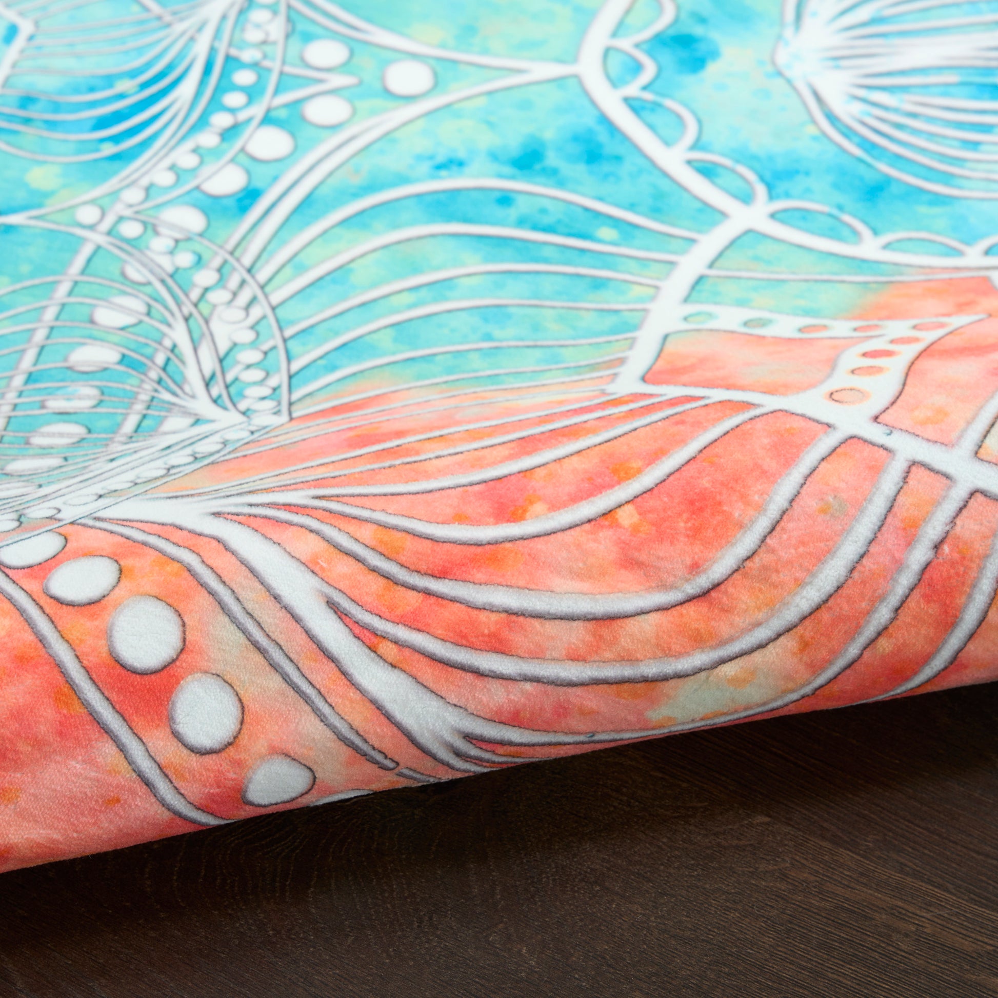 Close-up fabric with blue-teal gradient, pink edge, white metallic lines forming abstract floral/mandala pattern on wood.