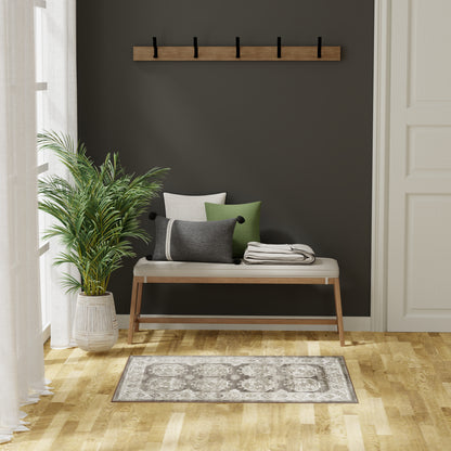 Wooden entry bench with beige cushion and green/gray pillows on a dark wall, coat rack above, potted plant, patterned rug.
