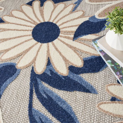 Floral area rug featuring a large cream daisy with navy center and blue leaves on beige woven backdrop; plant nearby.