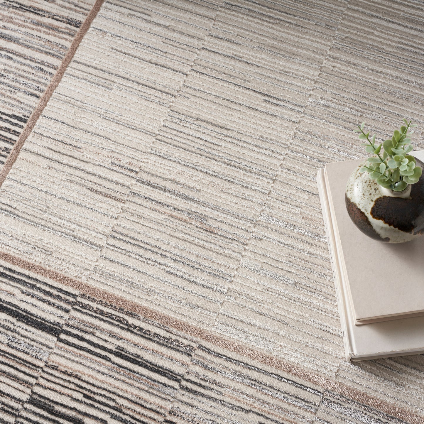 Neutral striped area rug in beige, cream and taupe tones with a small succulent in a speckled pot atop a book stack.