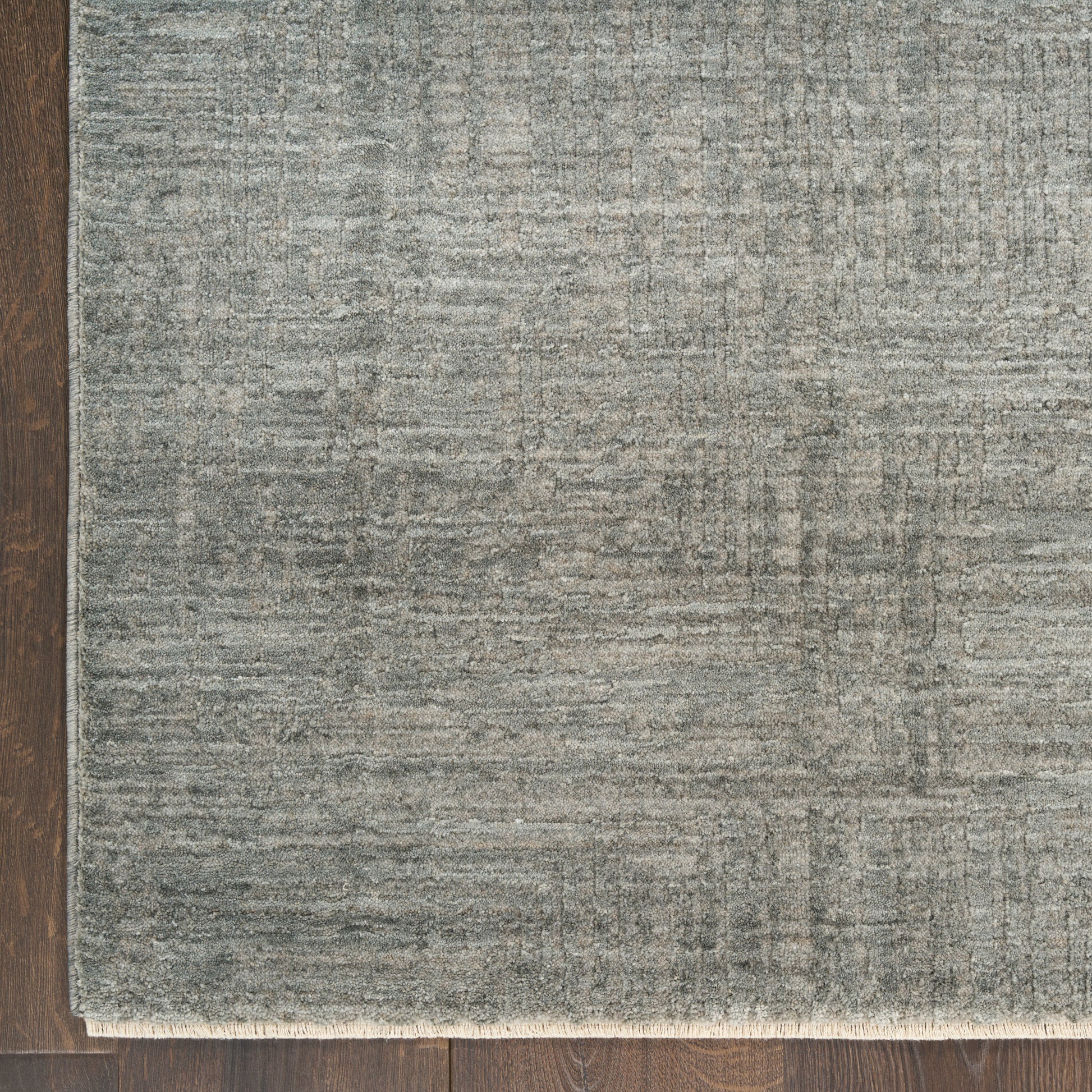 Rectangular gray-blue rug with subtle textured pattern and light fringe along bottom edge on dark wood floor.