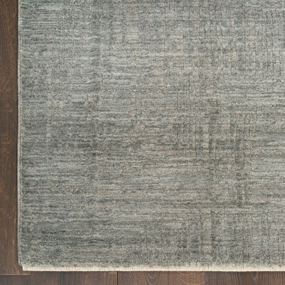 Rectangular gray-blue rug with subtle textured pattern and light fringe along bottom edge on dark wood floor.