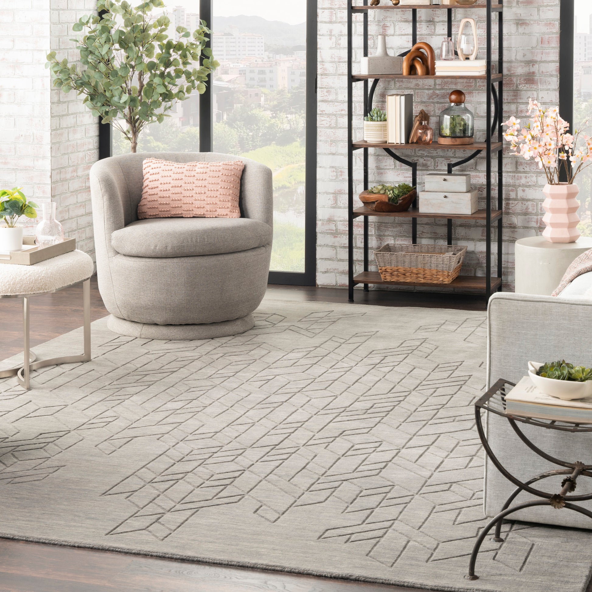 Bright modern living room with round gray lounge chair, pink cushion, geometric rug, black metal shelving & plants, windows.