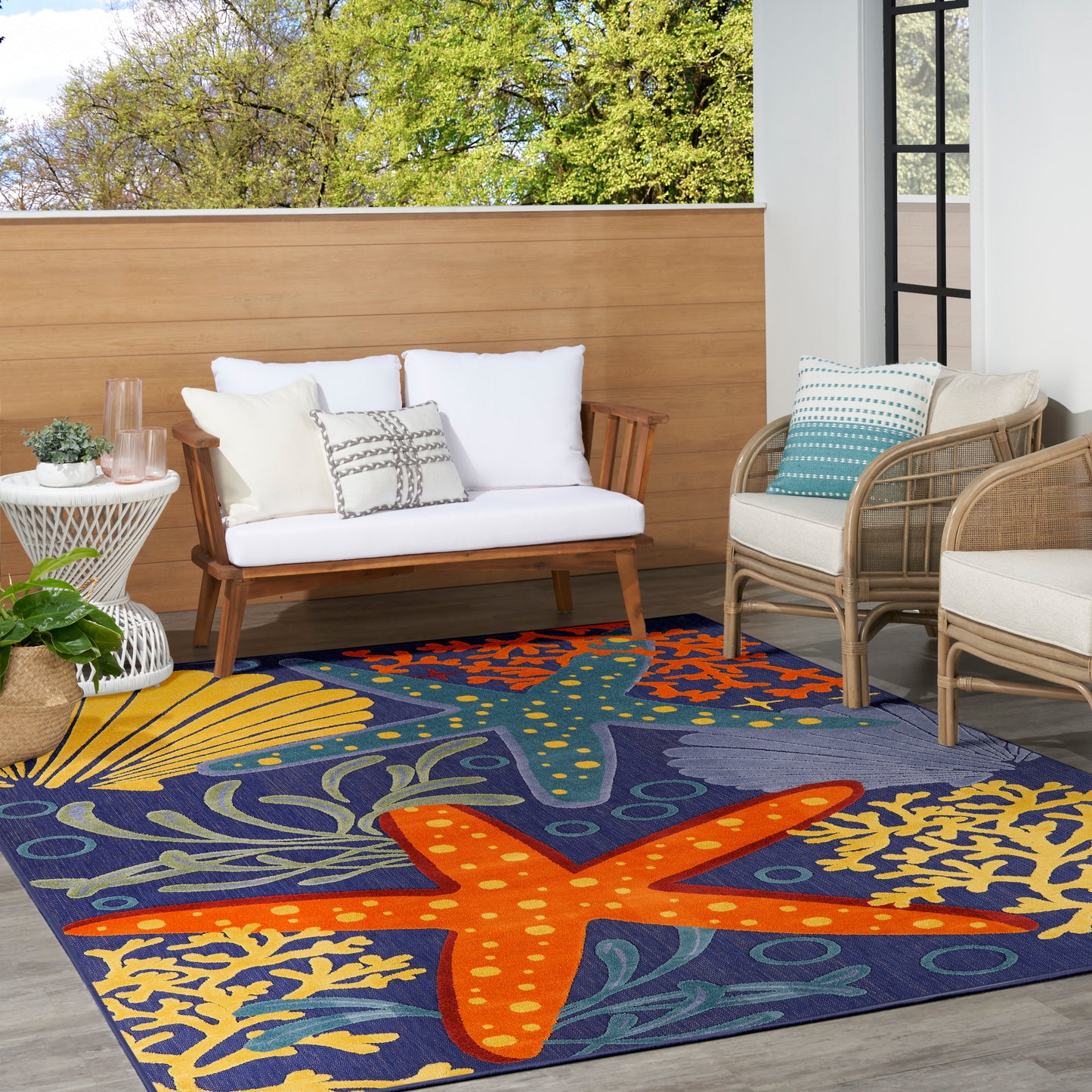 Outdoor space with a bold blue rug featuring orange starfish and seaweed, white-cushioned wooden loveseat, and wicker chairs.