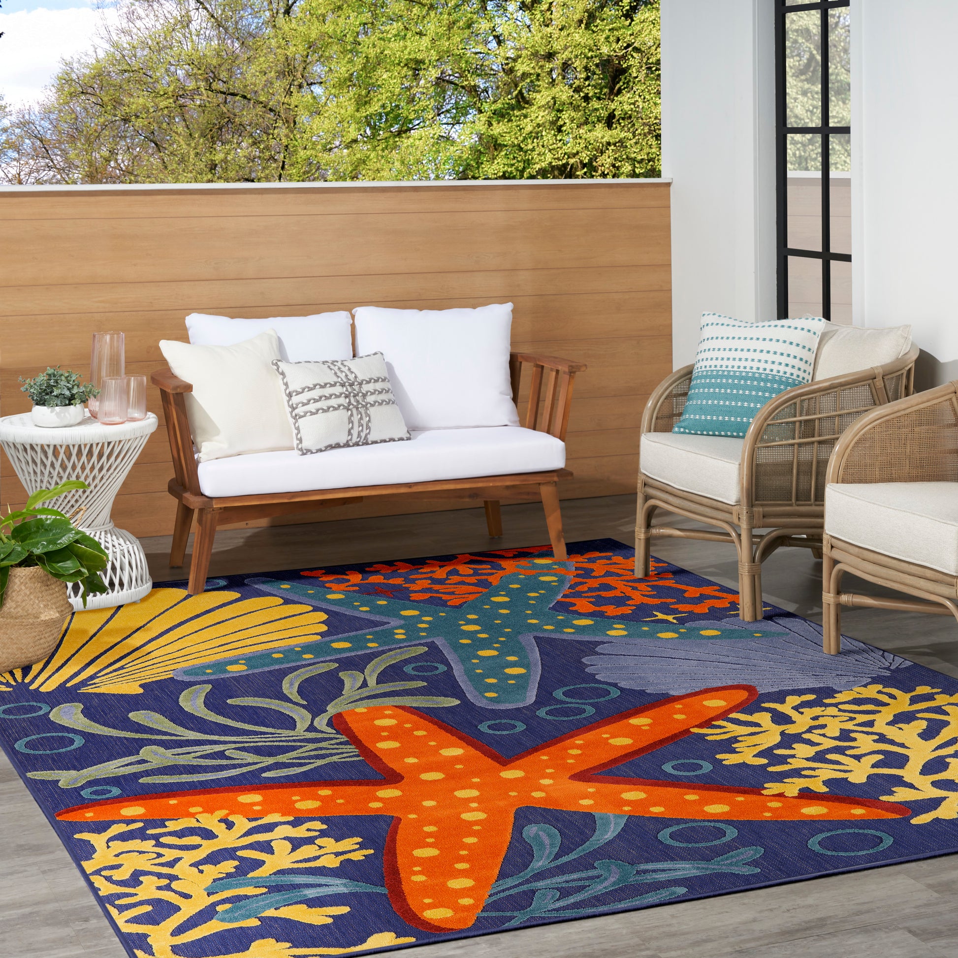Outdoor space with a bold blue rug featuring orange starfish and seaweed, white-cushioned wooden loveseat, and wicker chairs.