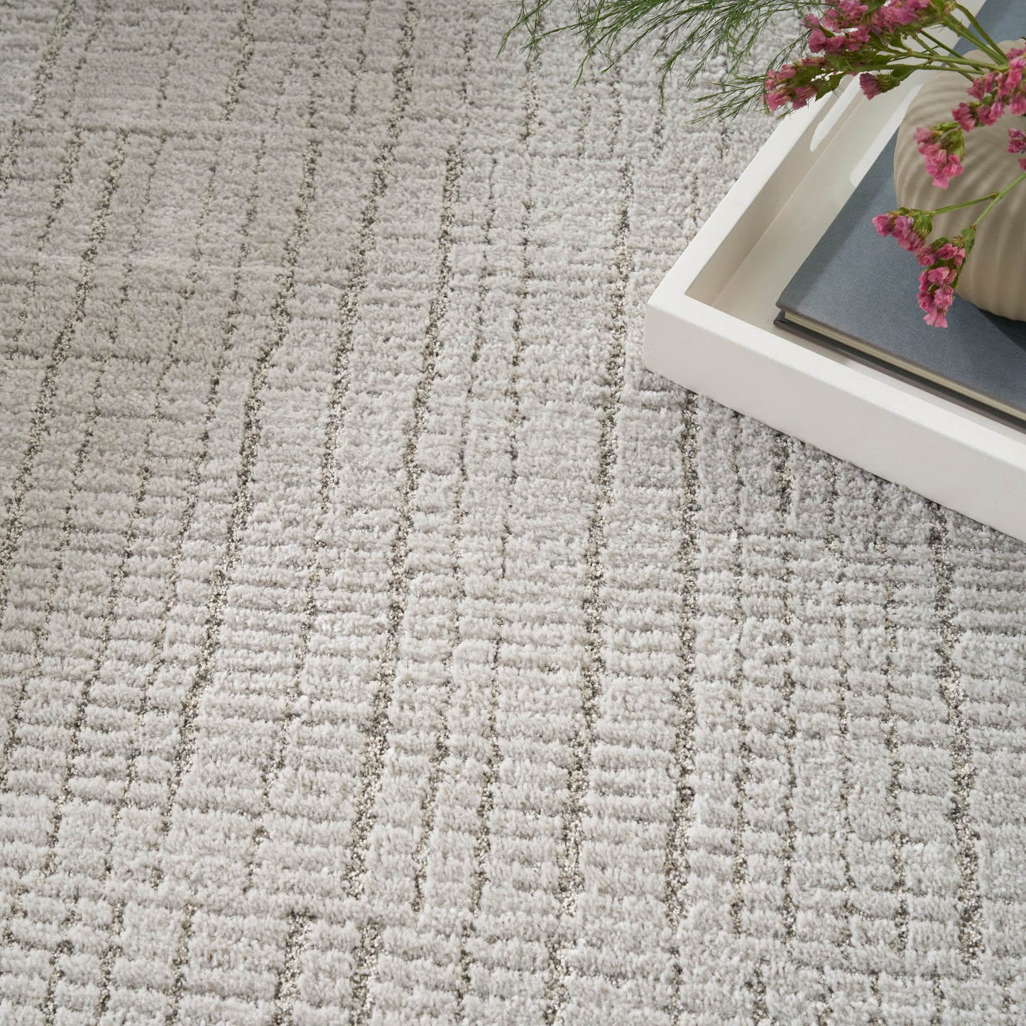 Soft gray-beige textured area rug with a subtle geometric grid; white tray with books and pink flowers in corner.