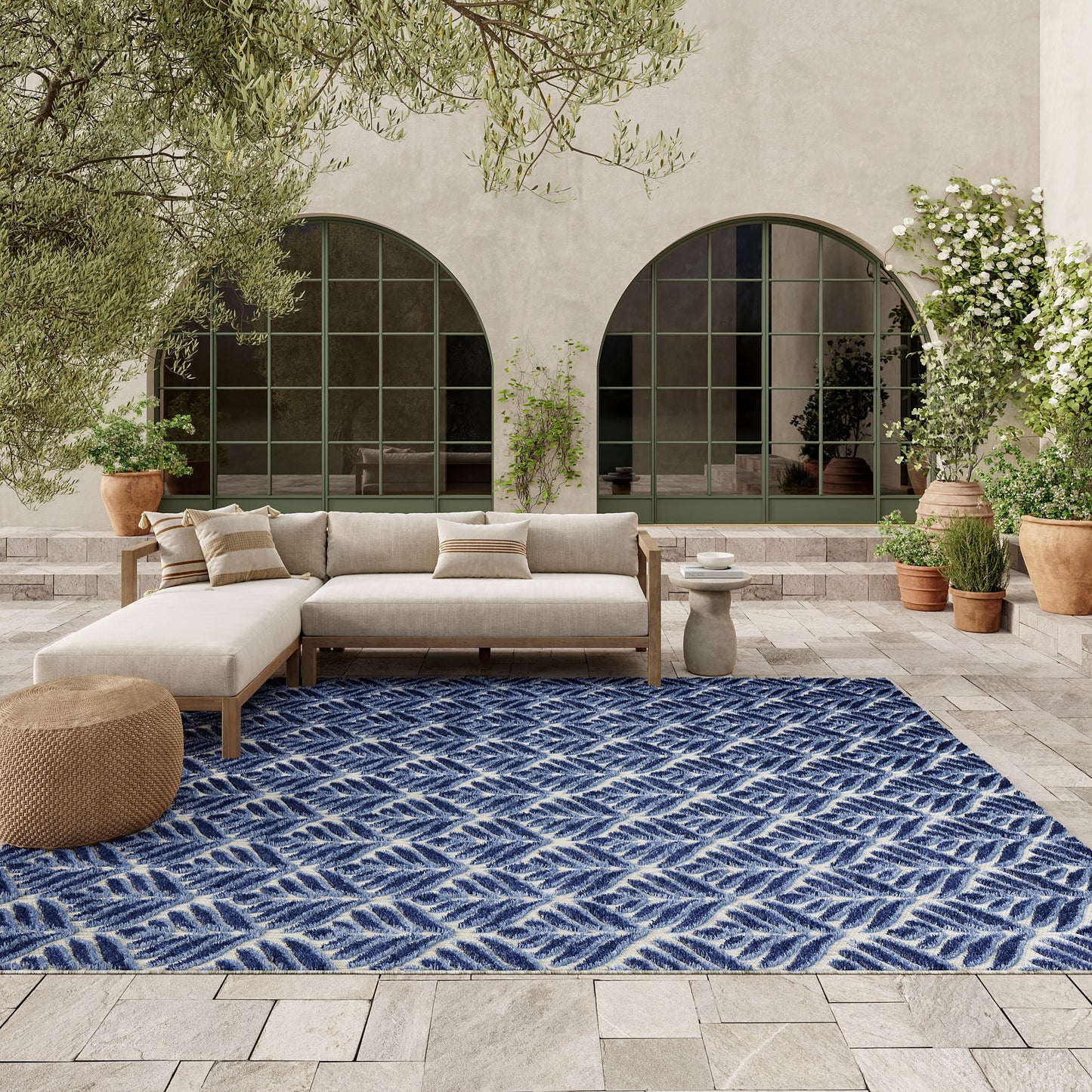 Outdoor lounge on a stone patio with a navy blue geometric rug, beige sectional sofa, pouf and potted greenery.