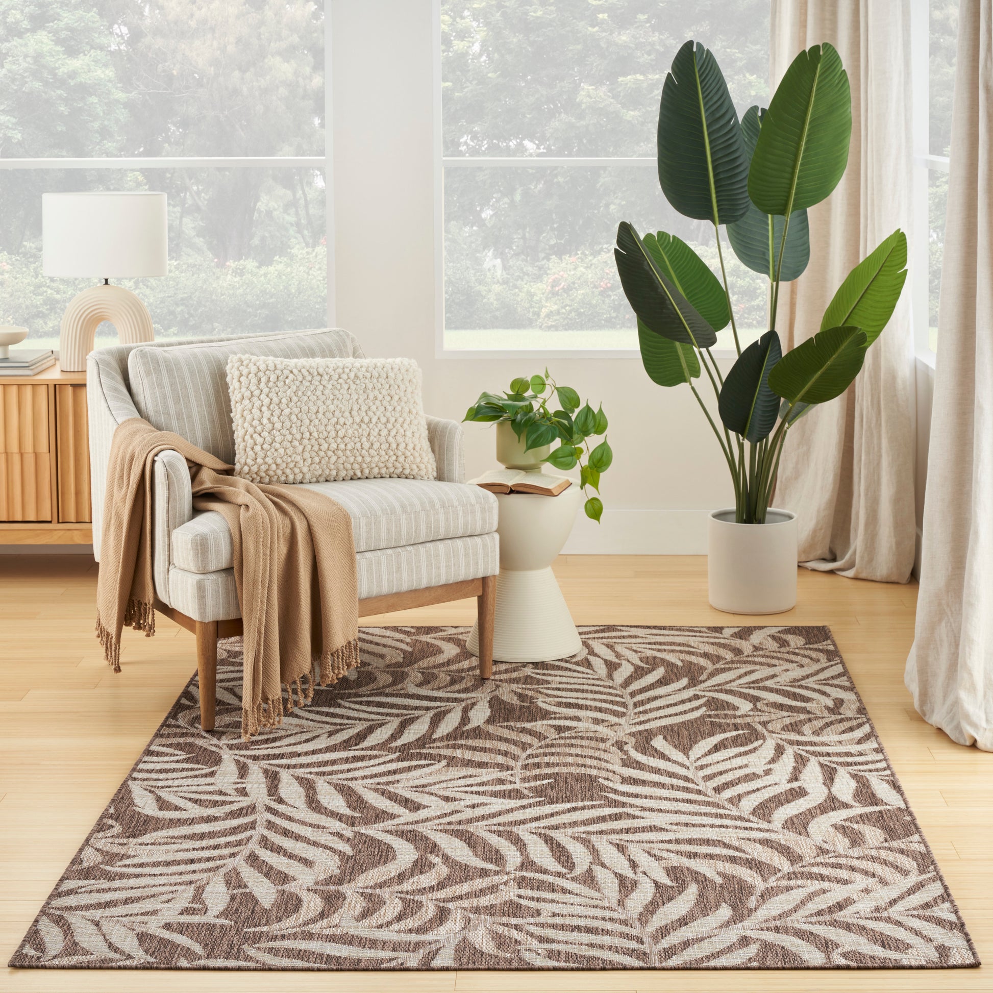 Cozy living room with beige striped armchair, tan throw, cream pillow, wood sideboard with lamp, tall leafy plant, and a brown leaf-pattern rug.