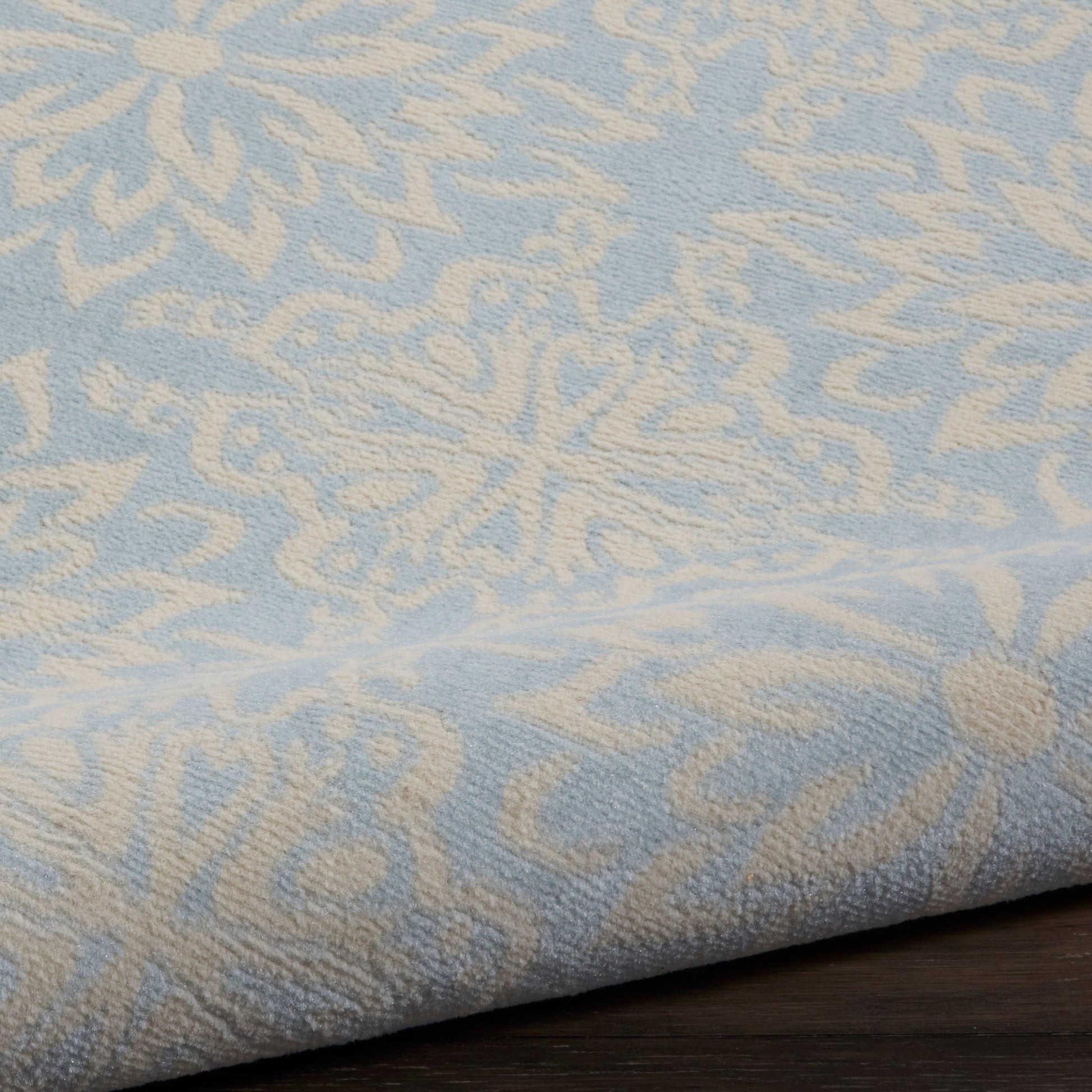 Light blue throw blanket with cream damask pattern, jacquard weave, folded edge on dark wood surface.