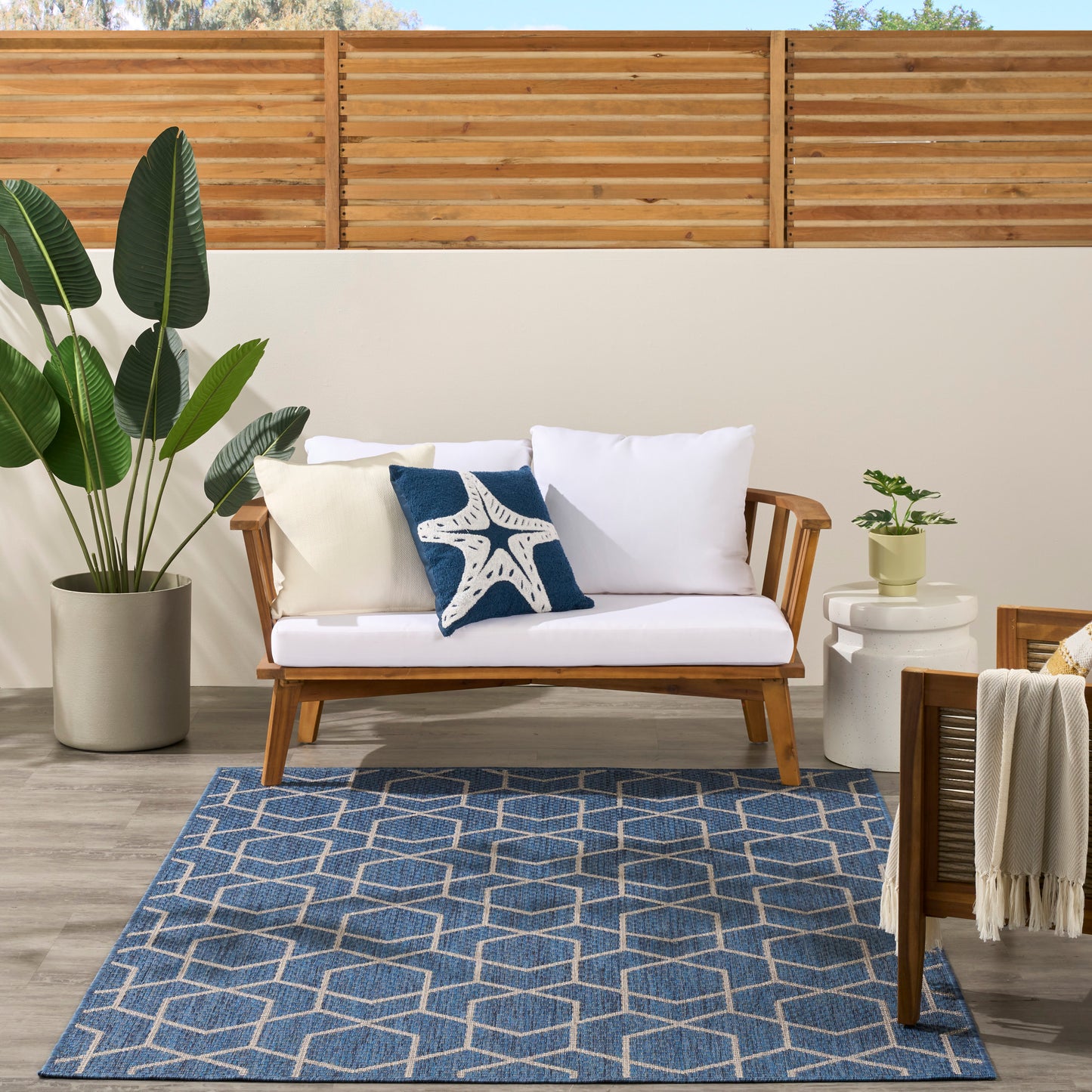 Outdoor patio with wooden-framed sofa, white cushions, starfish pillow, navy geometric rug, large potted plant.