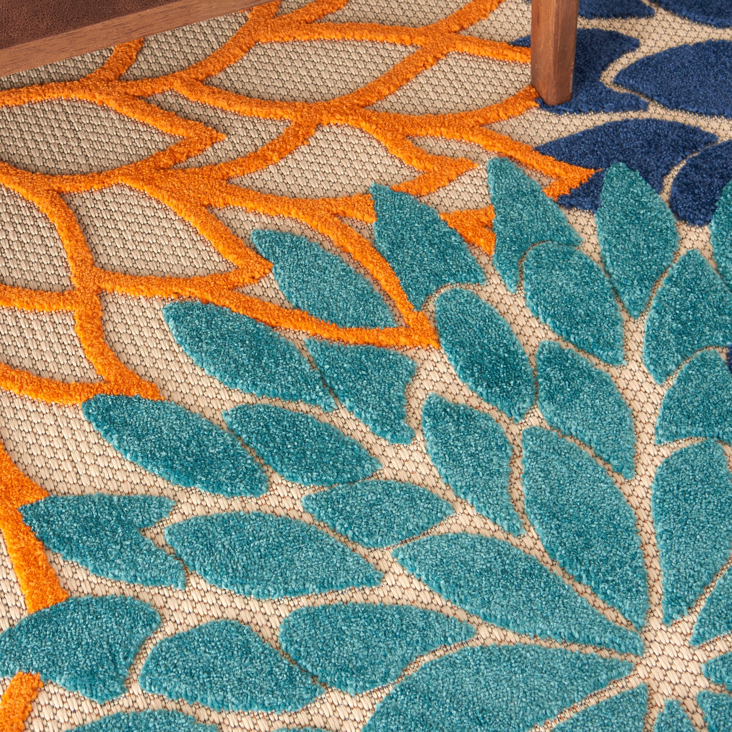 Close-up of teal leaf-pattern rug with orange lattice details on beige background; wooden table leg visible.