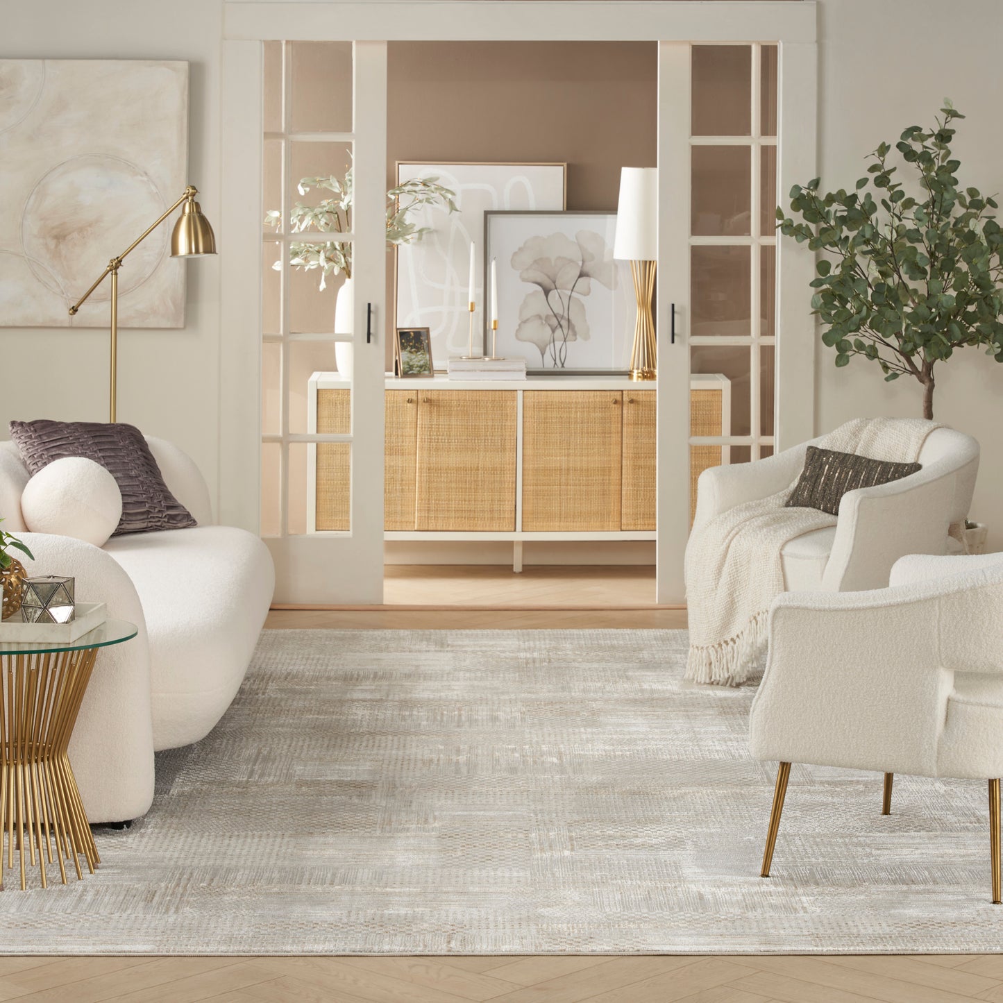 Bright beige and white living room with curved white sofas, gold lamp, glass side table, neutral rug, and open French doors.