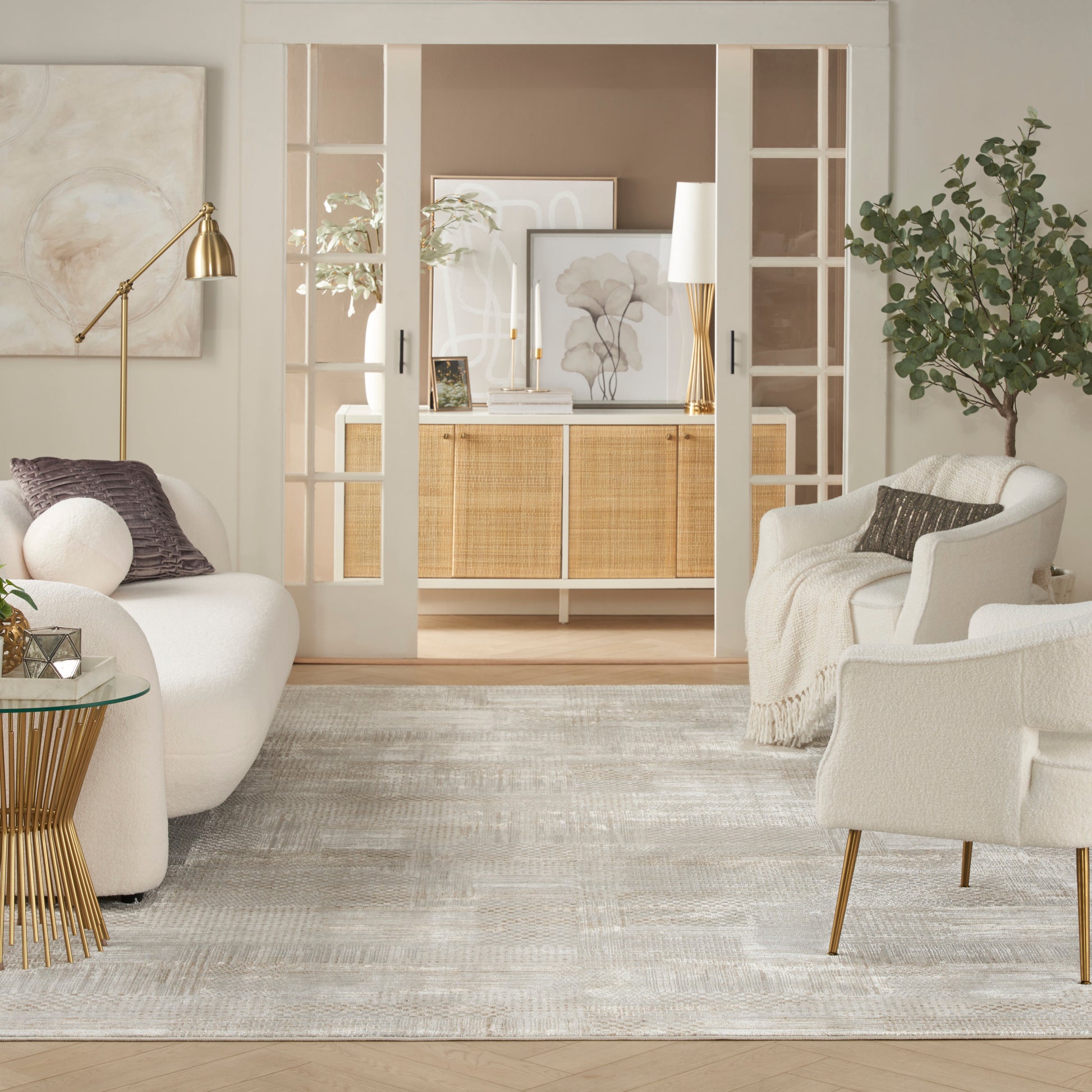 Bright beige and white living room with curved white sofas, gold lamp, glass side table, neutral rug, and open French doors.