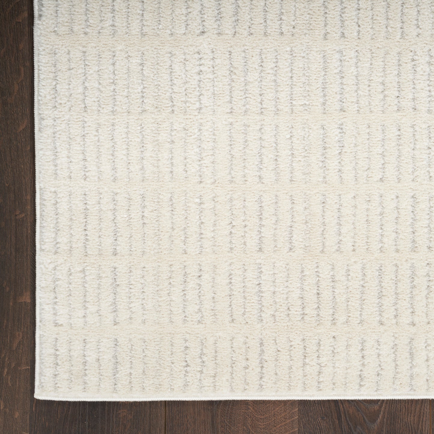Beige textured area rug with subtle raised vertical stripe pattern, cream tones, white edge binding, on dark hardwood floor.