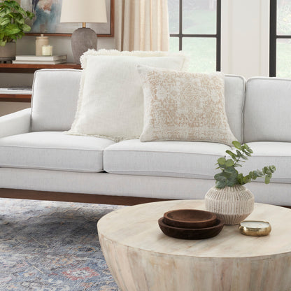 Light gray sofa with white fluffy and beige patterned cushions; round light wood coffee table with vase and bowls in a neutral living room.