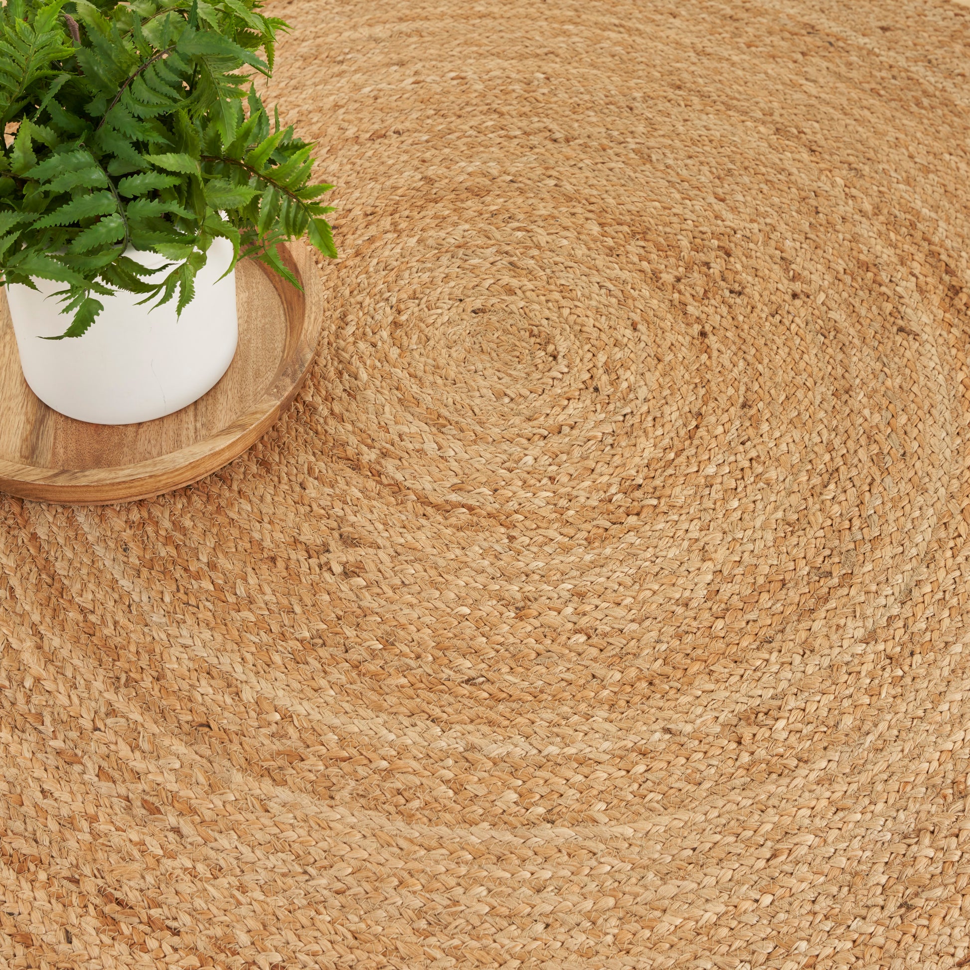 Round natural jute rug with a woven spiral pattern, white ceramic pot on a wooden tray with a green leafy plant.