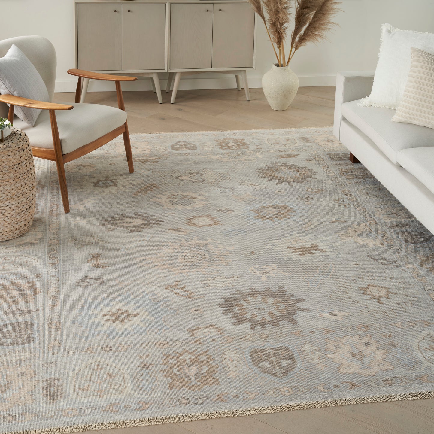 Light living room with a pale floral rug, mid-century armchair, woven side table, pale sofa, and pampas grass vase.