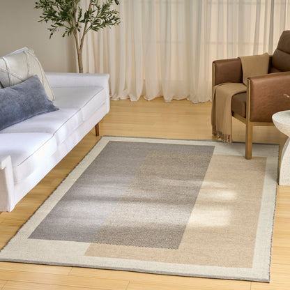 Neutral area rug with beige border & grey blocks on light wood floor in a bright living room with white sofa & brown chairs.