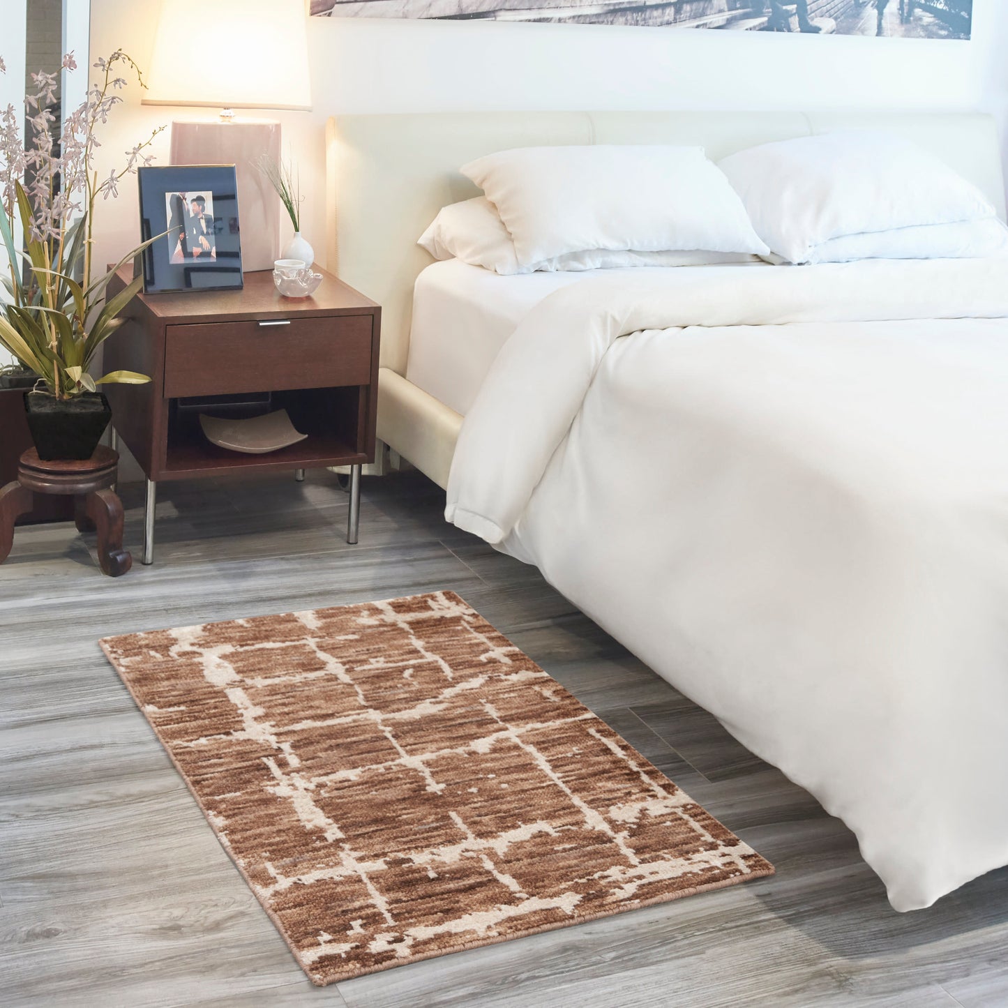 White bedding on a modern bed with dark wood nightstand, lamp, plant, framed photo; brown geometric rug on gray wood floor.