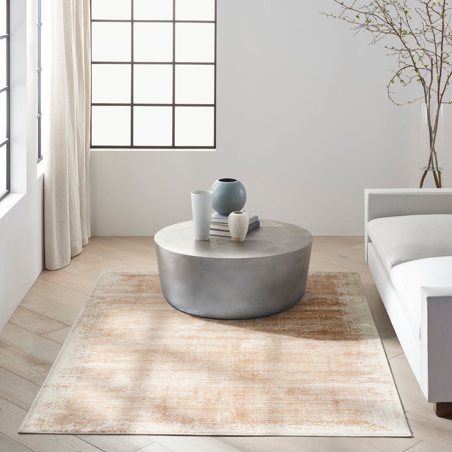 Minimalist living room with round concrete coffee table, blue vases, beige rug, light wood floor, large grid window and sofa.