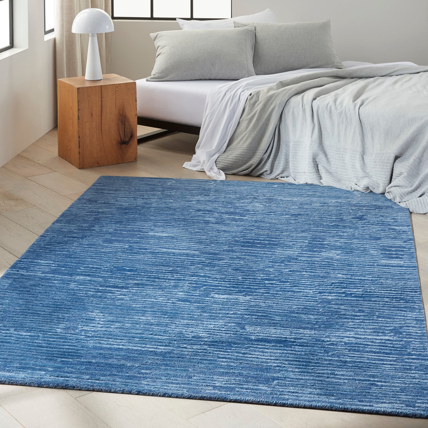 Blue textured rectangular area rug in a modern bedroom with light wood floor, gray bedding, and a wooden nightstand.