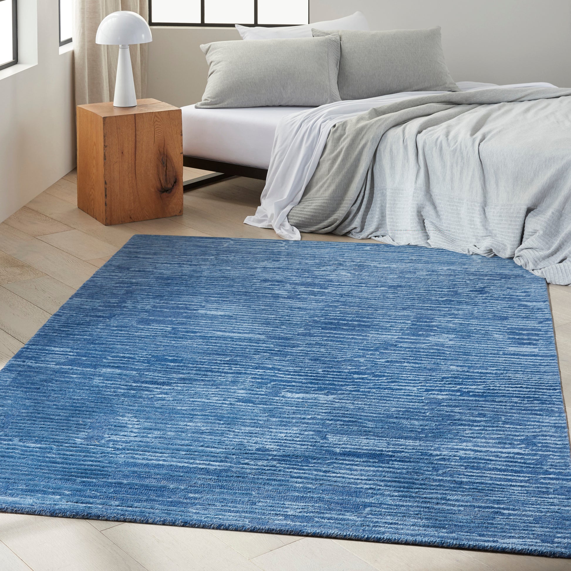 Blue textured rectangular area rug in a modern bedroom with light wood floor, gray bedding, and a wooden nightstand.