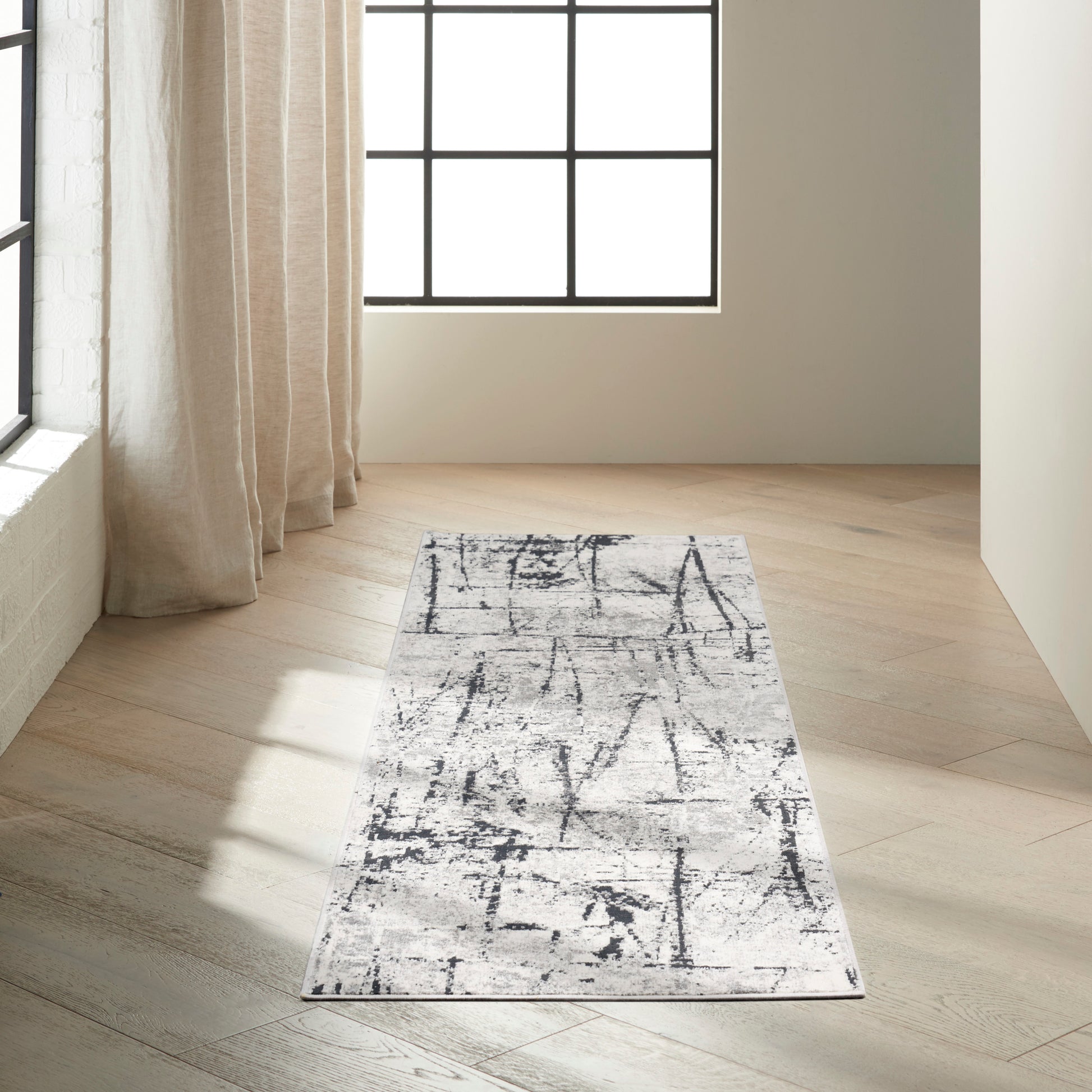 Light grey abstract-pattern rug runner on pale wood floor in a bright minimalist room with beige curtains.