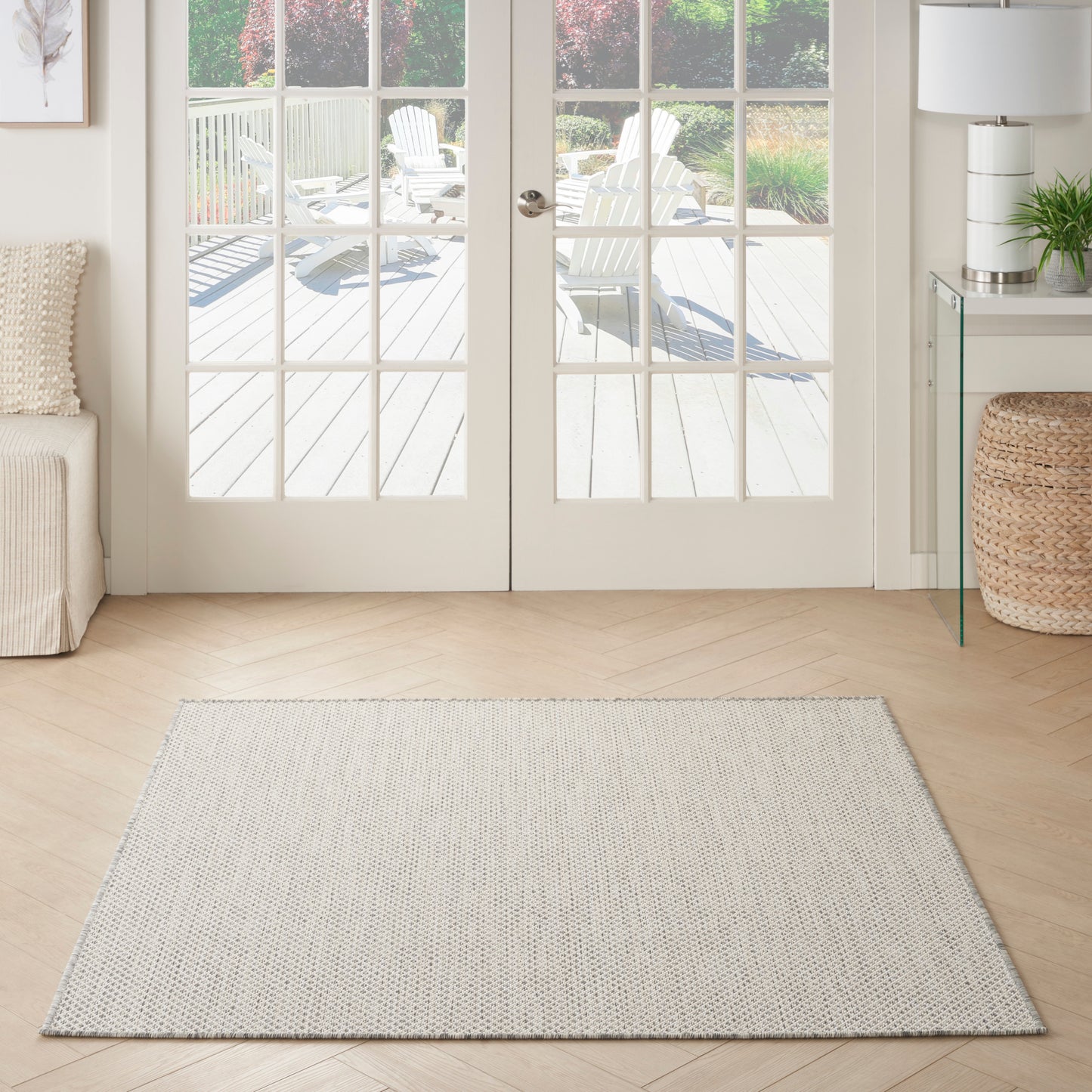 Rectangular light gray woven area rug on pale wood floor in a bright living room with French doors opening to a deck.