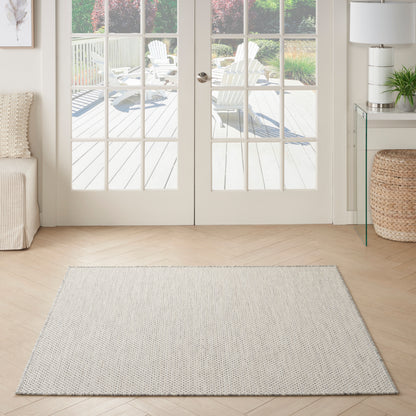 Rectangular light gray woven area rug on pale wood floor in a bright living room with French doors opening to a deck.