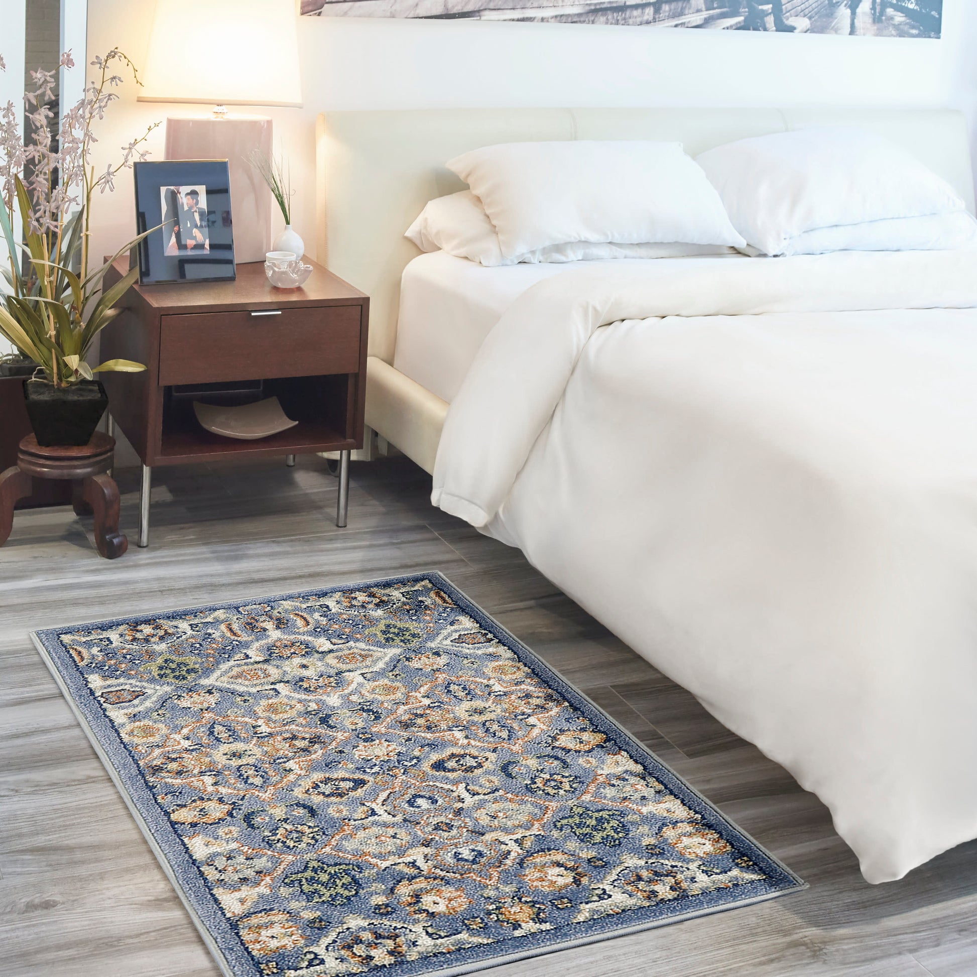 White bed with cream bedding, wood nightstand, lamp, plant, framed photo; blue and ivory bohemian rug on gray floor, 3' x 5'.