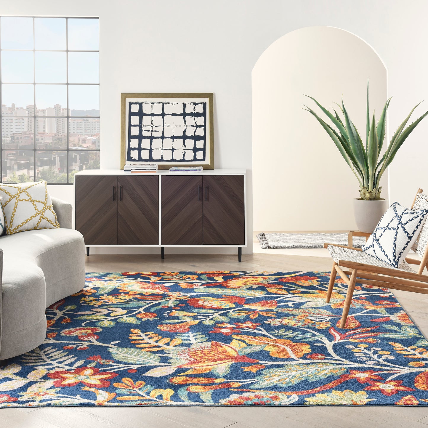 Bright living room with blue floral rug, gray sofa, dark wood mid-century sideboard, abstract art, wicker chair and plant.