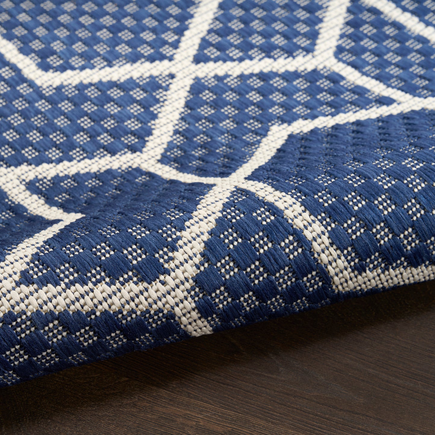 Blue and white geometric woven rug with diamond grid pattern and chunky texture, partially rolled on dark wood floor.