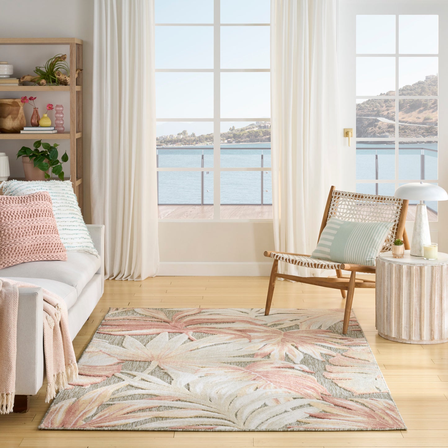 Bright coastal living room with large windows, pastel tropical rug, wicker chair, sofa with pink throw and striped cushions.