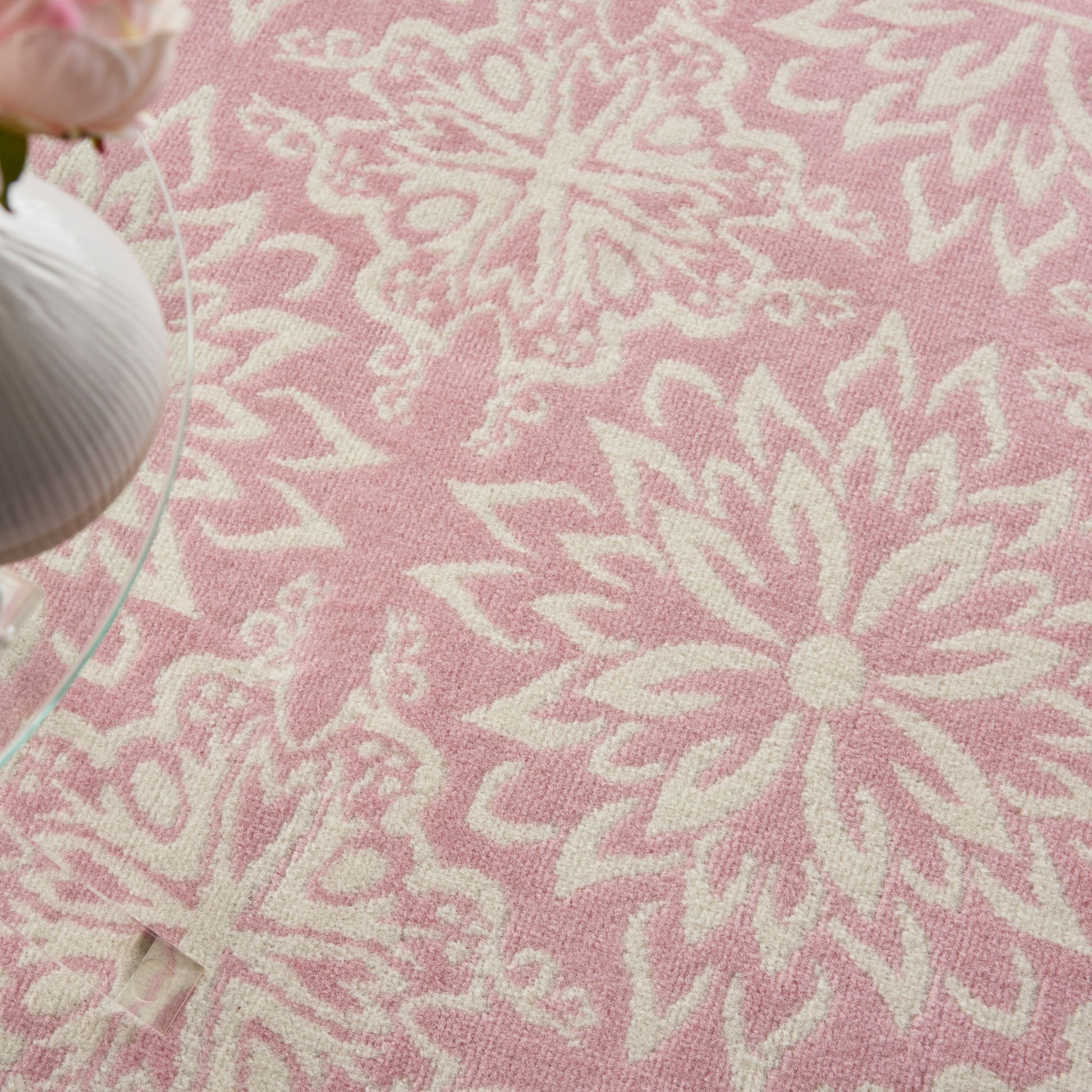 Soft pink rug with white floral/mandala patterns; round glass coffee table edge visible.