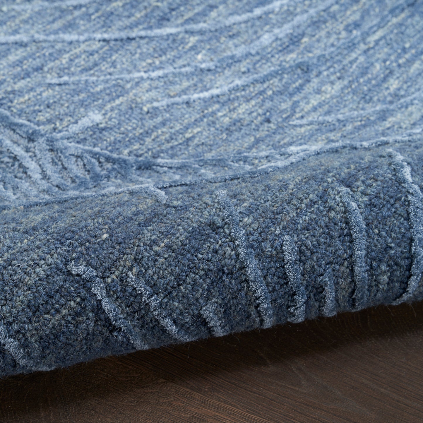 Blue plush rug with thick pile and subtle striped pattern, rolled edge resting on dark wood floor.