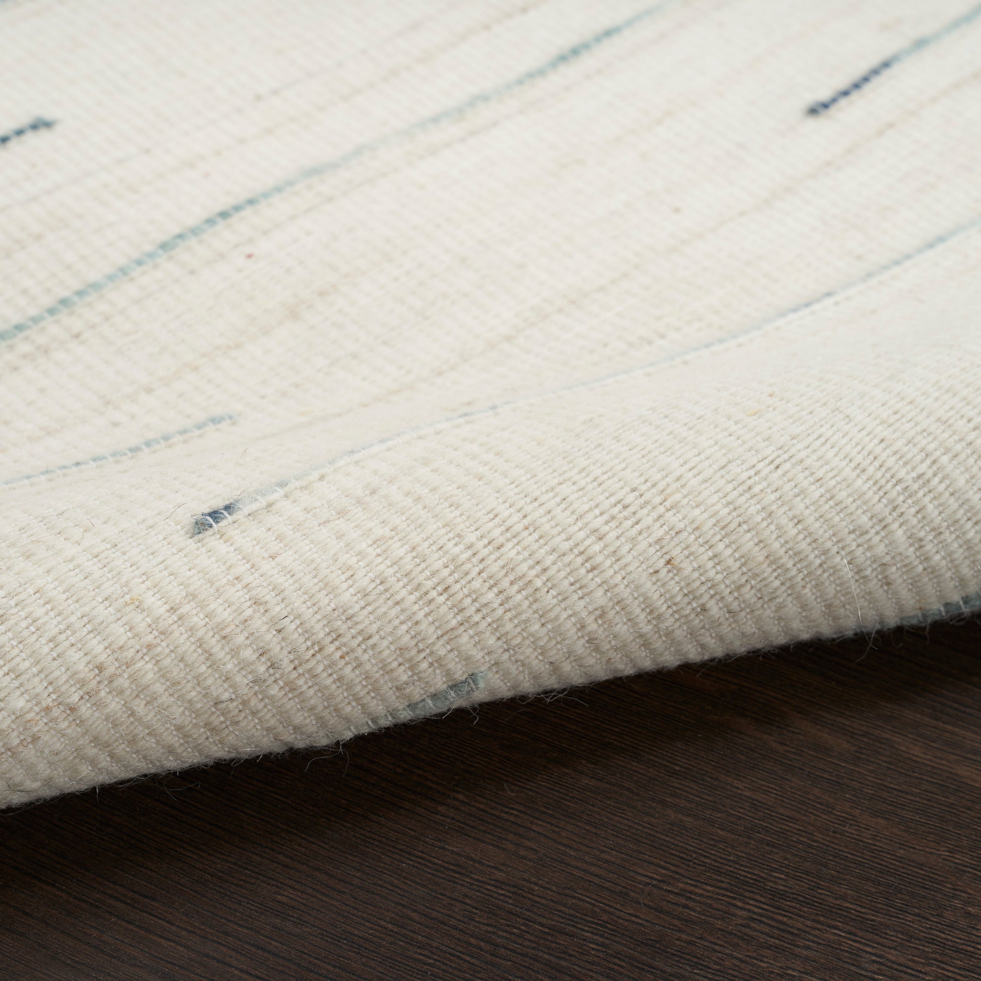 Cream woven rug with subtle light blue flecks, rolled edge on dark wooden floor.