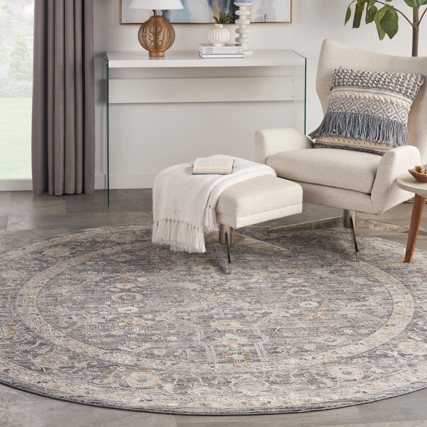 Circular grey oriental rug with beige medallions under a cream lounge chair, ottoman and glass console table.