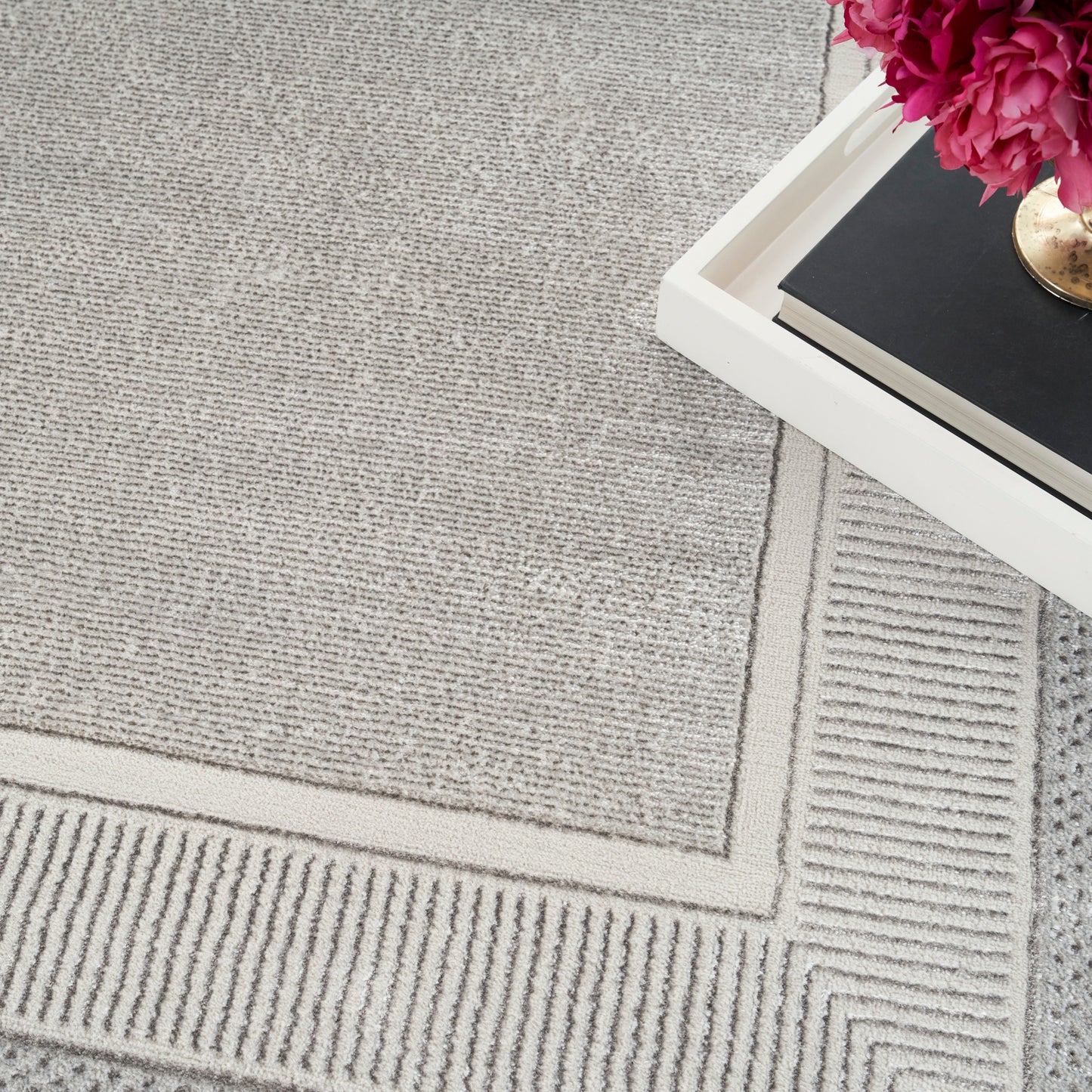 Light gray textured rug with a geometric border, white tray on top right holding a black book and pink flowers.